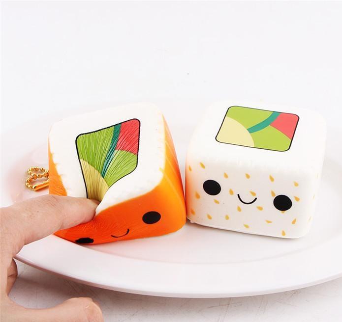 Two squishies that resemble sushi, the orange one is being squished by a finger. The other one is white with yellow spots and green and pink on top