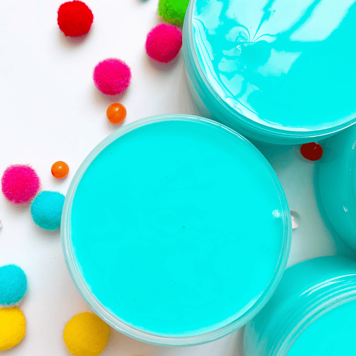 Several blue glossy slimes in their containers viewed from the top with multicoloured pompons in the background.