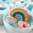 Close up to a  white and blue cloud slime decorated with a charm of a colorful rainbow with two clouds and a small pink heart on the right. The slimes if out of its container.