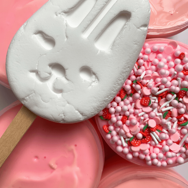 White rabbit clay popsicle with several glossy slimes in the background and one pink glossy slime with many pink and white round beads, white sprinkles and red strawberry slices sprinkles. 